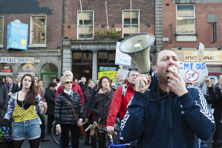 Protester speaking into a megaphone at a street demonstration, surrounded by a crowd with signs in an urban setting.