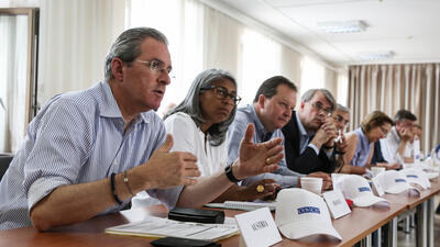 Several individuals, including men and women, sit at a conference table engaged in a discussion. The man in the foreground is speaking, gesturing with his hands, while others listen attentively. Small nameplates and white hats with the OSCE logo are visible on the table.