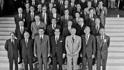 Black-and-white photograph showing the Foreign Ministers of the CSCE participating States, gathered for the first Helsinki meeting in 1973. The group is posed on a set of steps, with several rows of individuals dressed in formal suits and ties. In the background, other attendees and onlookers are visible, adding depth to the setting.