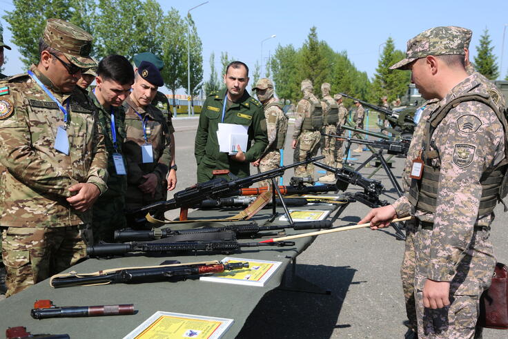 Military personnel examine displayed rifles at an outdoor exhibition, guided by an officer in camouflage uniform.