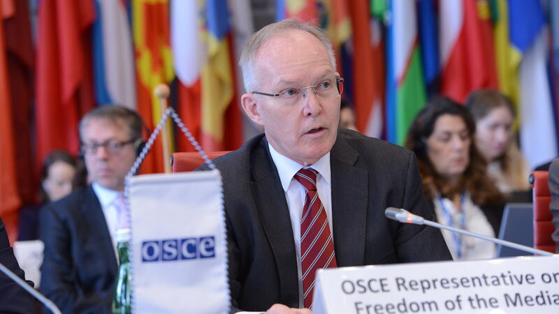SCE Representative on Freedom of the Media, Ambassador Jan Braathus, speaking to the OSCE Permanent Council, seated at a table with a nameplate in front of him and flags as well as listeners in the background.