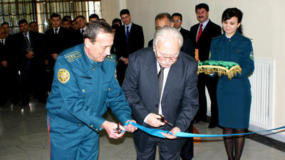 Two men cutting a ceremonial ribbon, surrounded by people, with a uniformed woman holding a decorative cloth.