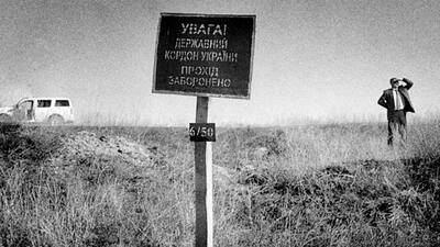 A man stands in a grassy field beside a sign, with a white SUV parked in the background.
