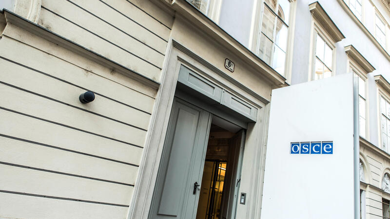 This image shows the entrance to the Organization for Security and Co-operation in Europe (OSCE). The building's facade is light-colored with horizontal grooves and rectangular windows that are symmetrically aligned. Above the entrance, the building's street number ("6") is visible on a small plaque. A white panel with the OSCE logo displayed is positioned near the doorway. The door is slightly open, offering a glimpse into the building's interior, which has glass doors inside.