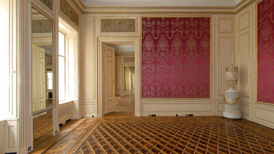 The image shows an elegant, historical interior. The room features intricate parquet flooring with a geometric pattern, ornate wall paneling in cream and gold, and rich red damask wallpaper. Large mirrors and windows reflect light, enhancing the sense of space. A decorative urn on a pedestal adds a classical touch. The open doorways lead to similarly adorned rooms, creating depth and continuity in the architectural design.