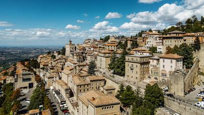 Hilltop town with tan buildings under a blue sky, surrounded by trees, and expansive landscape views.
