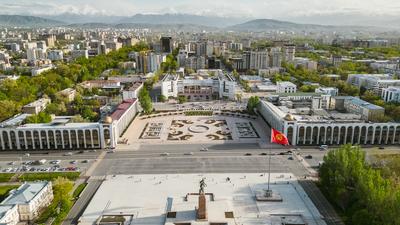 Urban landscape with large square, surrounded by greenery and buildings, mountains in the distance, red flag in foreground.