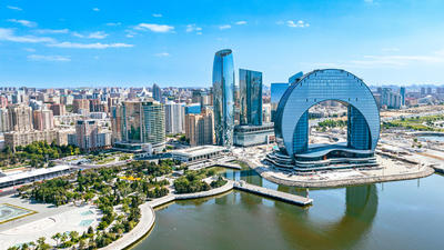Aerial view of a modern cityscape with a circular skyscraper by a river under a clear blue sky.