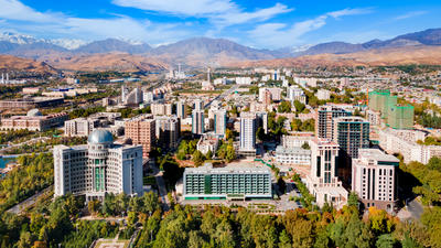 Aerial view of a cityscape with modern buildings, surrounded by green trees and mountains under a bright blue sky.