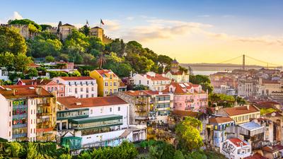 Colorful hillside homes in Lisbon with trees and a castle atop, a bridge in the background at sunset.