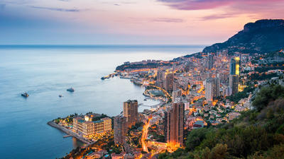 Coastal city at dusk, illuminated buildings line the shore, with mountains in the background and a colorful sky overhead.