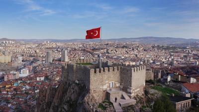 Aerial view of a historic stone castle with a Turkish flag, set against a sprawling cityscape under a blue sky.