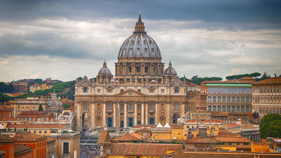 St. Peter's Basilica under a cloudy sky, surrounded by city buildings.