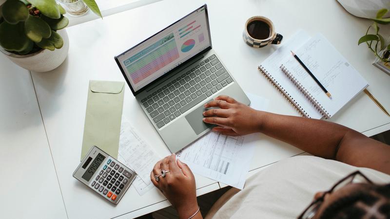 Person working at a desk with a laptop showing graphs, papers, a calculator, and a cup of coffee.
