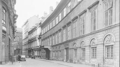 The black-and-white photo captures a historic Vienna street, where the OSCE Secretariat is located now. It features cobblestone roads, elegant multi-story buildings, and a façade with arched windows and decorative reliefs. A shop sign is visible, along with vintage cars and blurred pedestrians. The curved street adds depth, and the dark vignette suggests an old-fashioned camera.