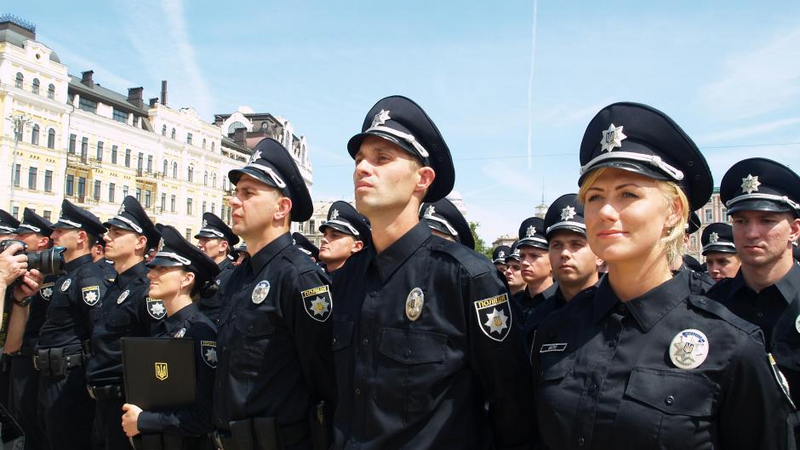 A group of uniformed police officers standing in formation outdoors. They are dressed in black uniforms with peaked caps featuring silver emblems. 